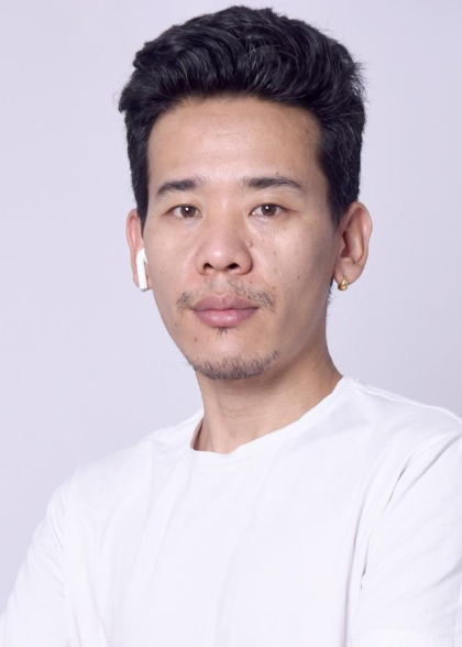 A young man with short dark hair, wearing a white shirt, a white wireless earbud in his right ear, a small earring in his left earlobe, and a slight goatee, posing against a light purple background.