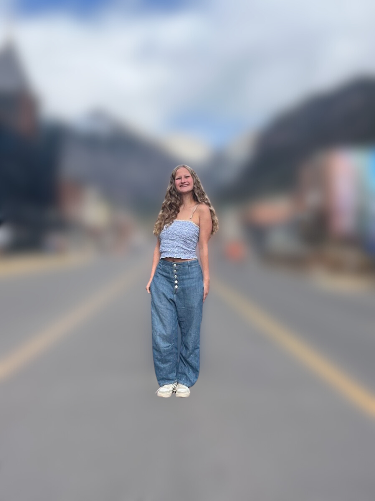 A young woman with long, wavy hair wearing a blue floral tank top, high-waisted blue jeans, and white sneakers standing in the middle of a blurred road with mountains and buildings in the background.