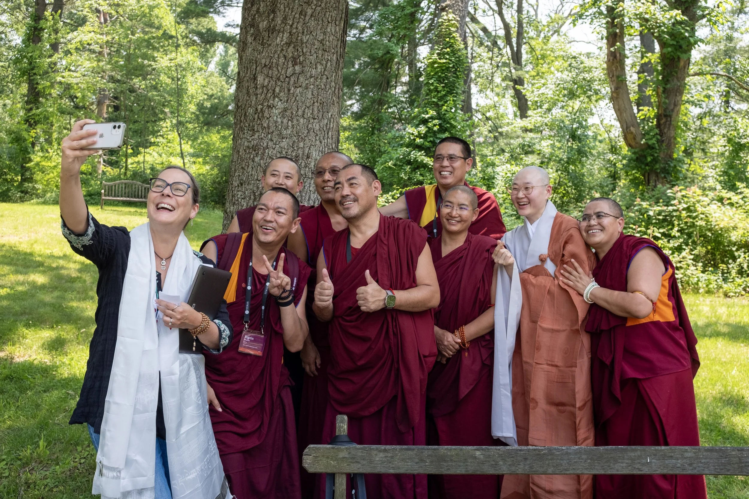 A group of people, including monks in maroon robes and women, taking a selfie outdoors in a lush green park with large trees.