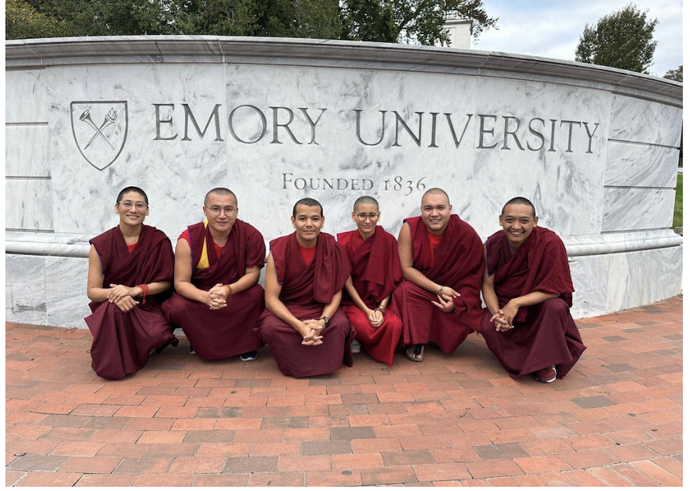 Six Buddhist monks in maroon robes pose in front of an Emory University sign.