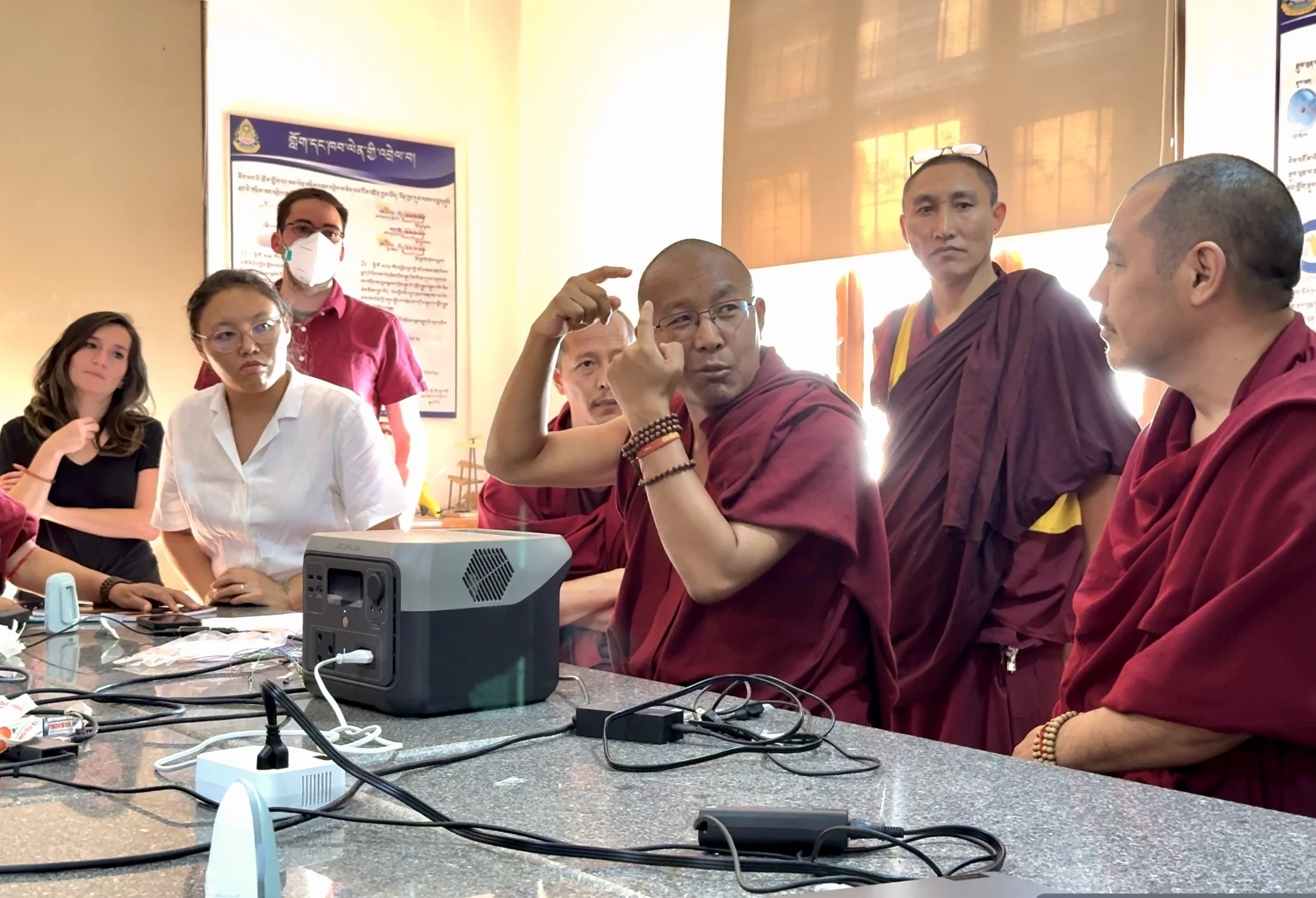 Group of people, including Buddhist monks and women, sitting and discussing around a table with electronic devices in a meeting room.