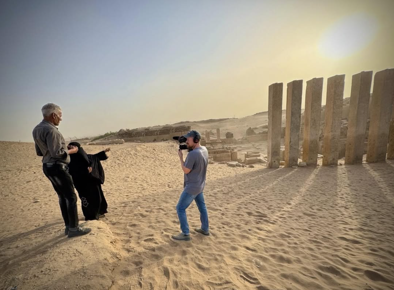Film crew interviewing a woman in a black abaya at a sunlit ancient desert archaeological site with stone columns.