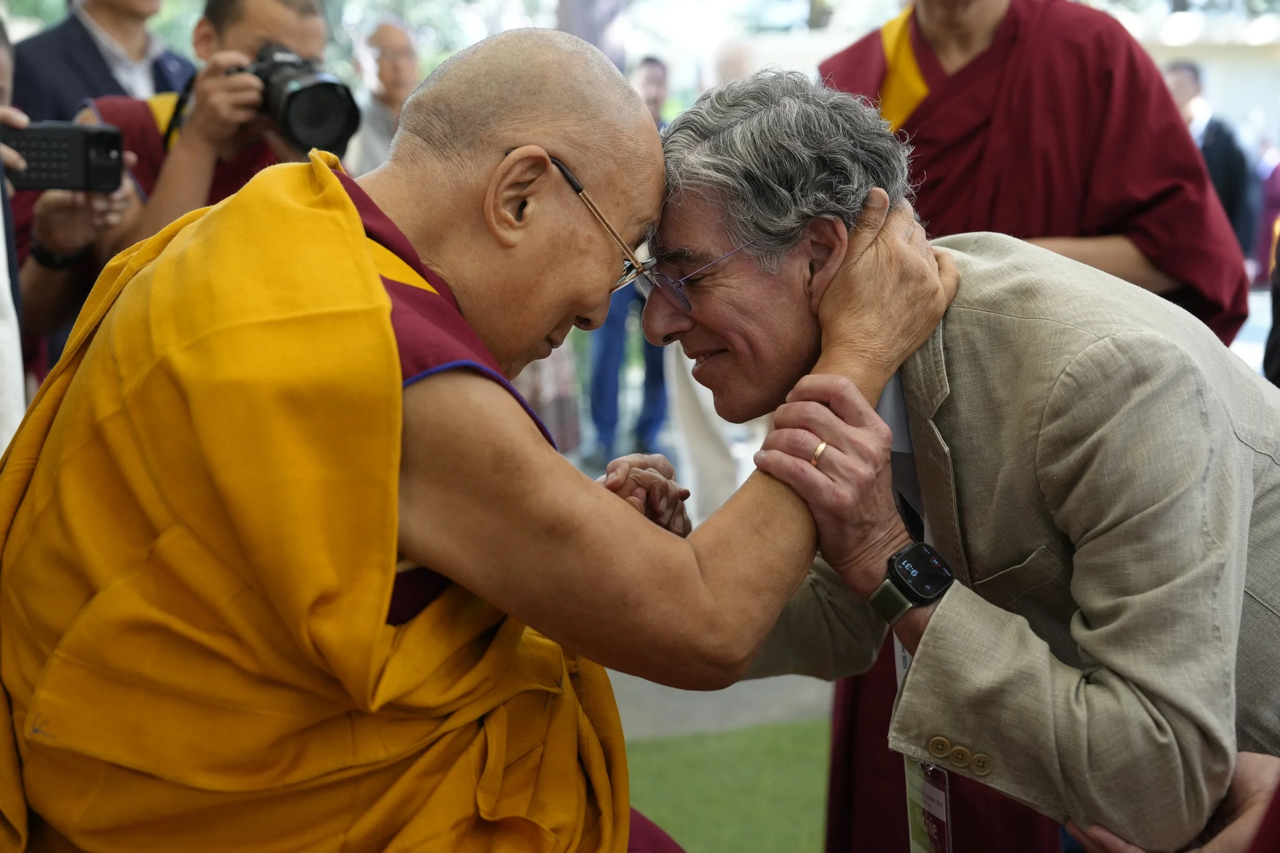 A Dalai Lama-like figure and a man are touching their foreheads together and holding hands, with eyes closed, in a gesture of compassion or connection. The man is wearing glasses and a beige suit, while the other person is dressed in traditional Tibe