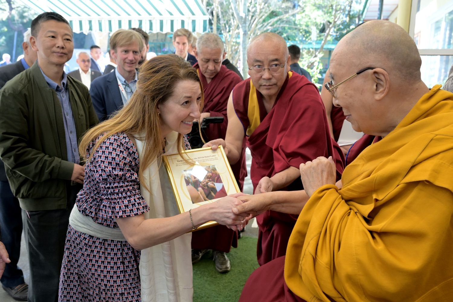 Woman presents a framed certificate to the Dalai Lama while Buddhist monks and attendees look on