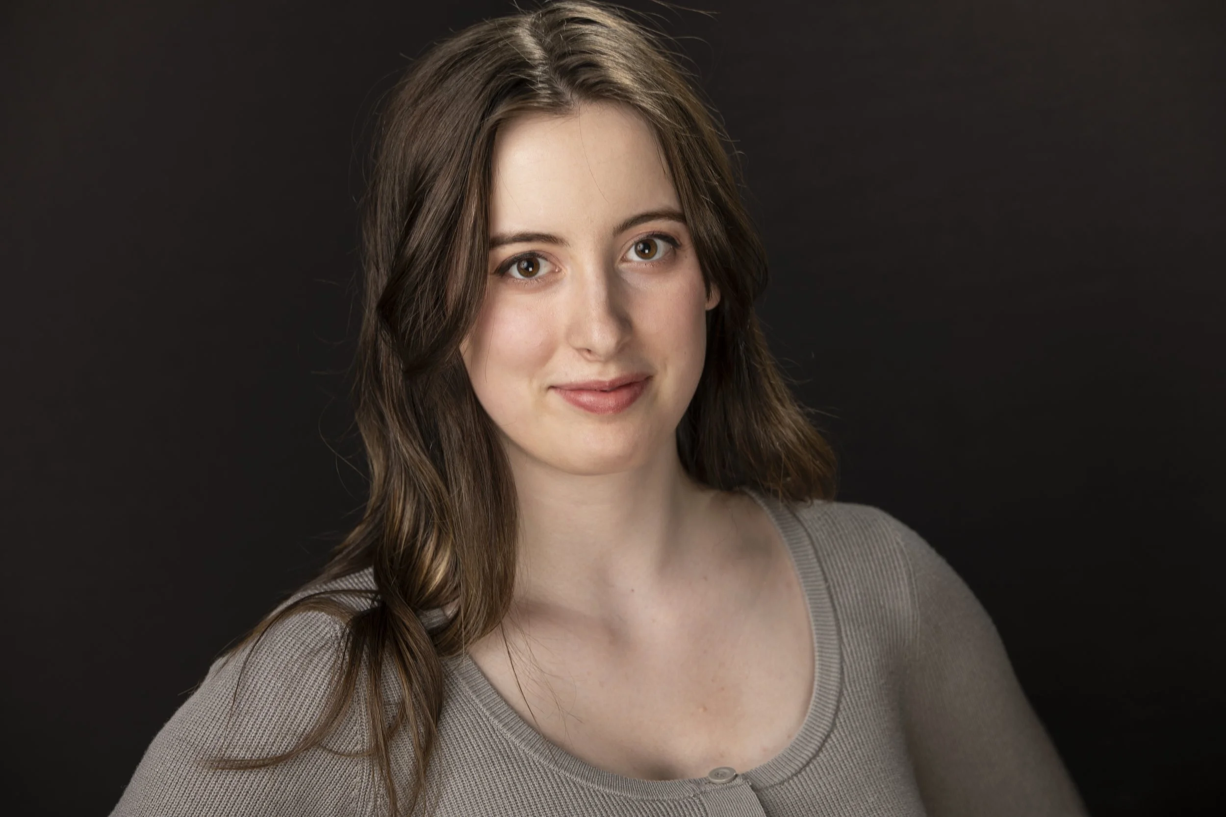 Portrait of a smiling woman with long brown hair wearing a gray sweater against a dark background