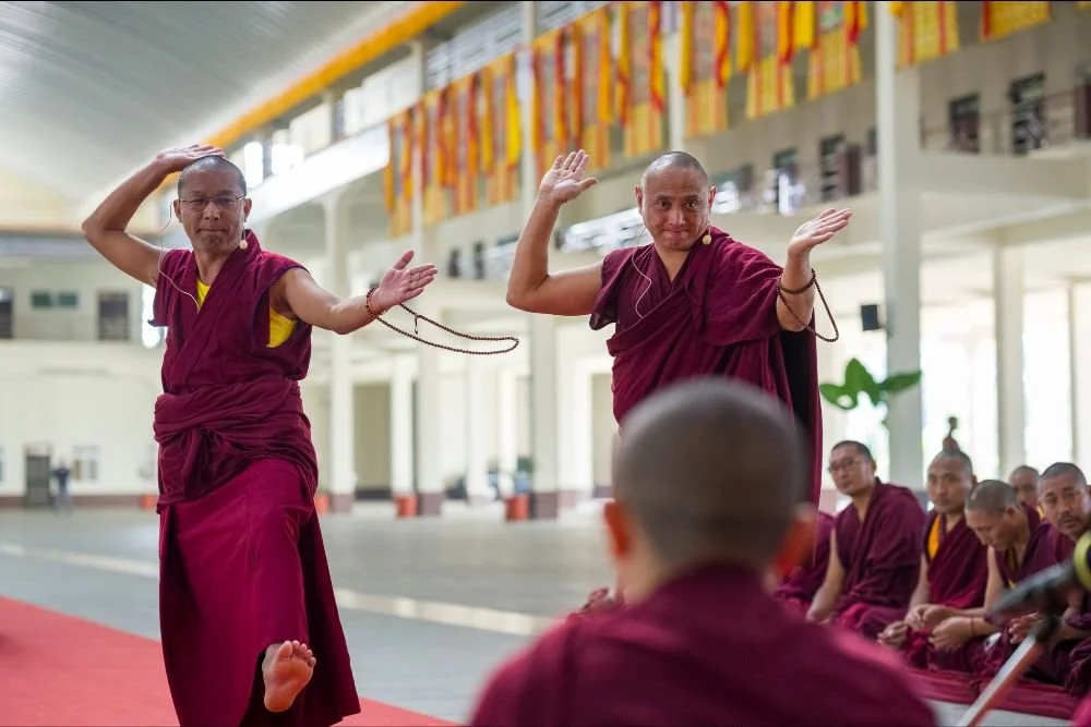 Buddhist monks performing a ritual in a spacious hall while seated monks watch.