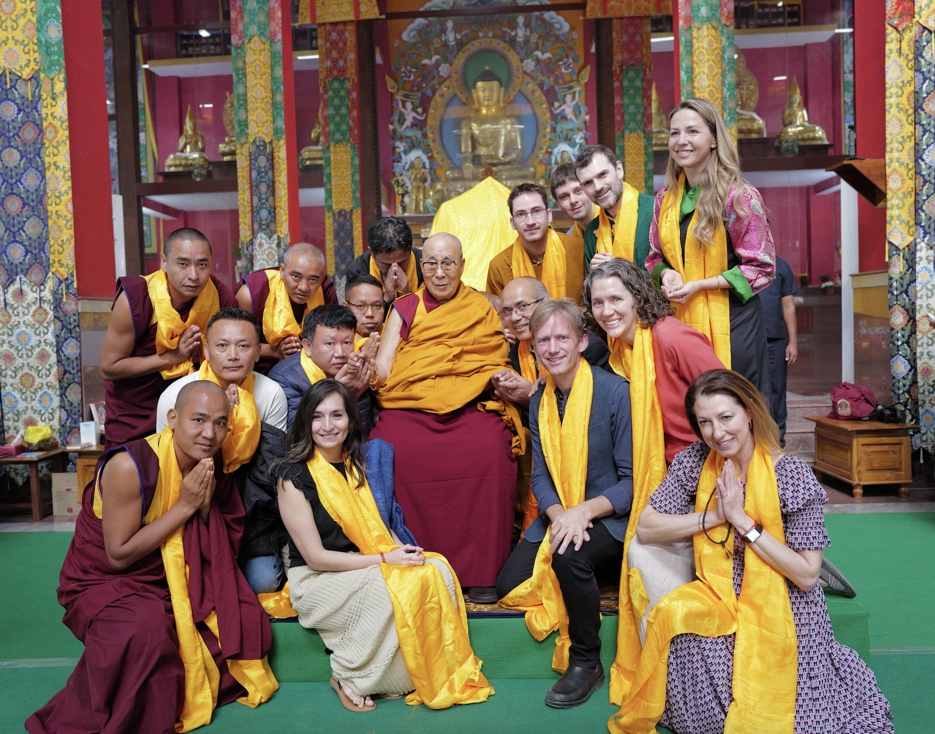 Group portrait of Buddhist monks and visitors wearing yellow ceremonial scarves inside a colorful Buddhist temple, with the Dalai Lama seated in the center.