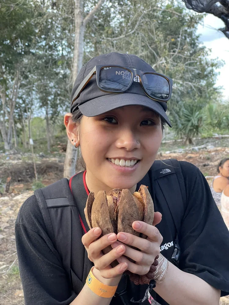 A woman smiling outdoors holding a dry, brown seed pod or flower with multiple segments. She is wearing a gray cap, sunglasses on her hat, and a black shirt with a backpack. The background shows trees and a forested area.