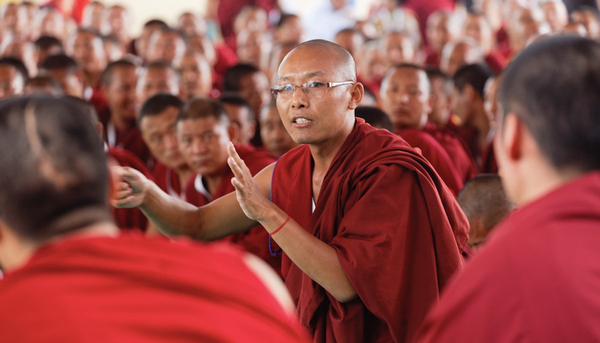 A Buddhist monk speaking to a group of monks dressed in red robes, in a large gathering.