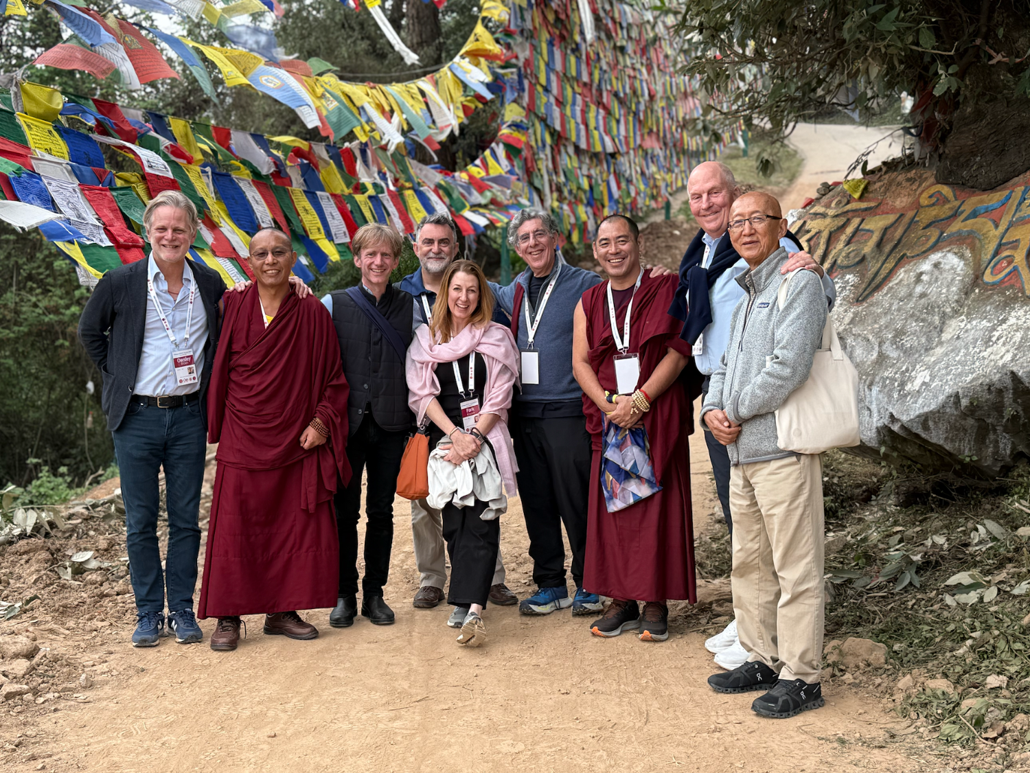 Group of nine people, including monks and tourists, pose together outdoors in front of colorful prayer flags at a cultural or religious site, with a dirt path and trees in the background.