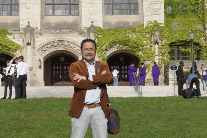 A man in a brown blazer and white shirt standing outdoors in front of a university building with ivy growing on the walls. Several people, including graduates in purple gowns, are visible in the background.