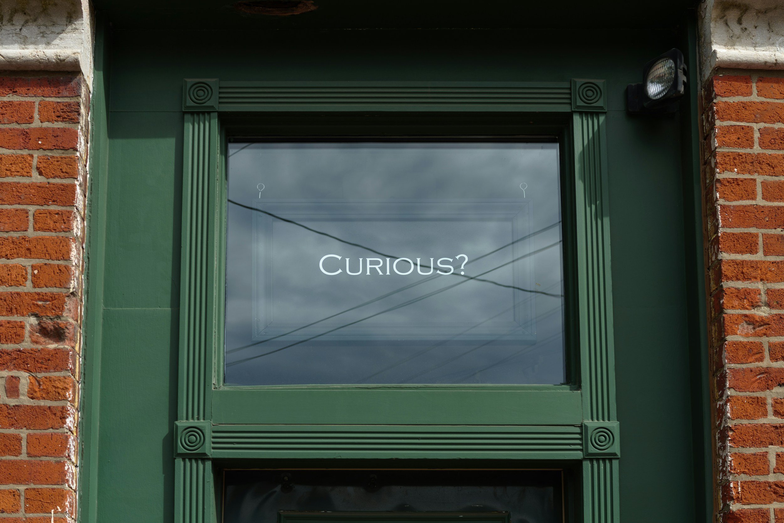 A window with green trim set into a brick building, with the word 'CURIOUS?' written across the glass.