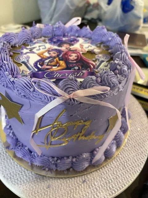 A birthday cake decorated with purple frosting and a picture of Disney's Elena of Avalor characters on top, with a "Happy Birthday" message and ribbons.