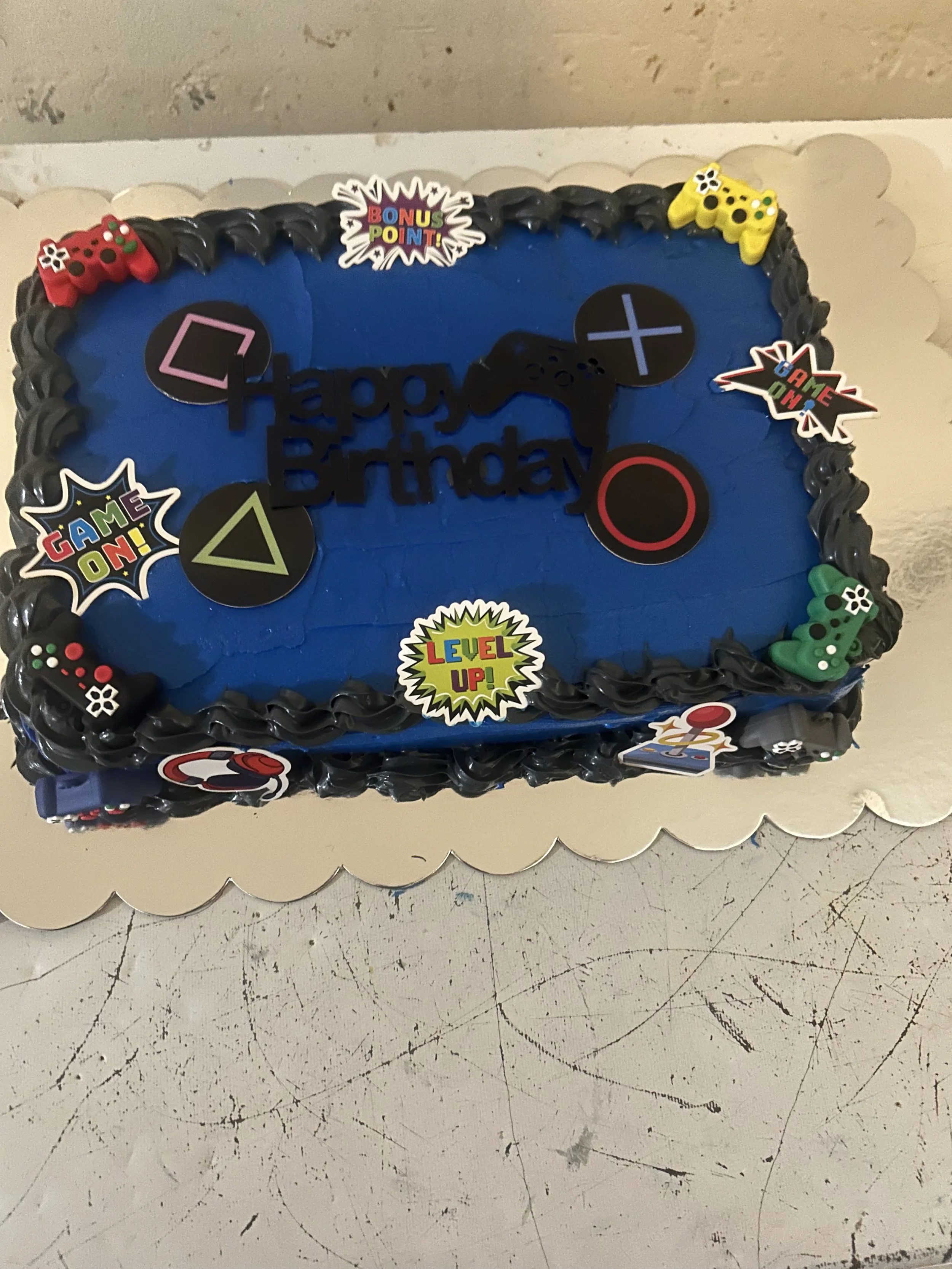 A rectangular cake with blue icing decorated with gaming-themed stickers, including game controllers, directional pad, and colorful labels such as 'Bonu Point,' 'Game On,' and 'Level Up.' The cake has a black frosting border and is placed on a white scalloped cake board.