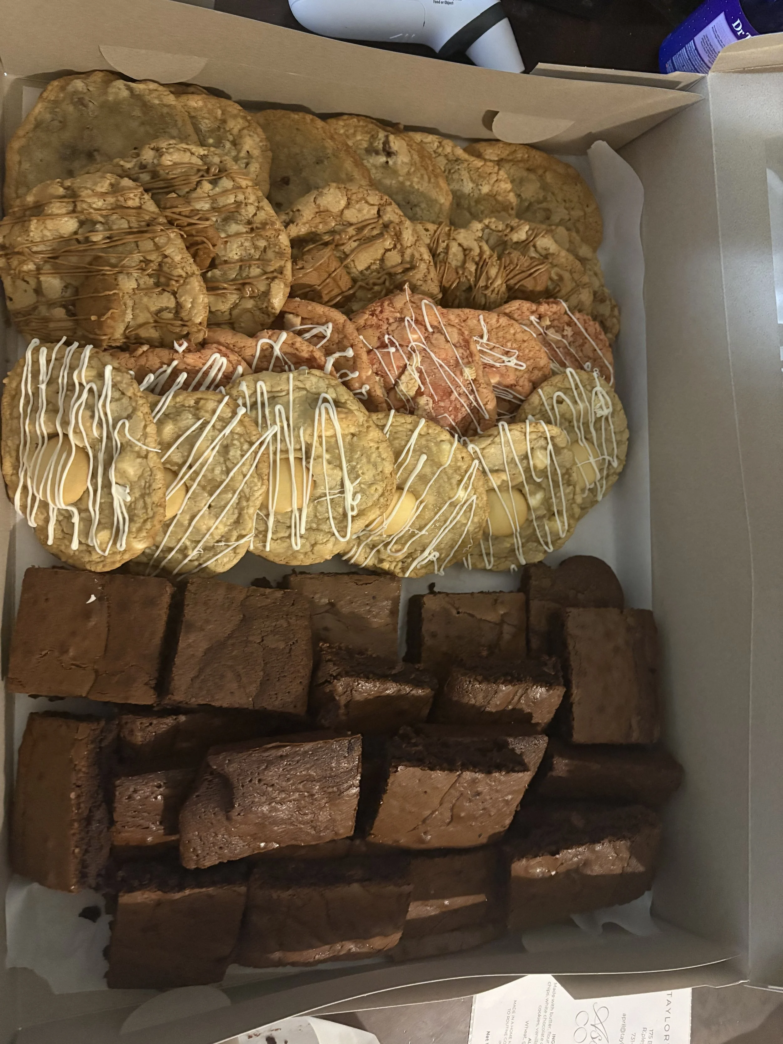 Box of assorted cookies on white paper, packaged in a cardboard box, with chocolate and white chocolate drizzles, stacked with brownies at the bottom.