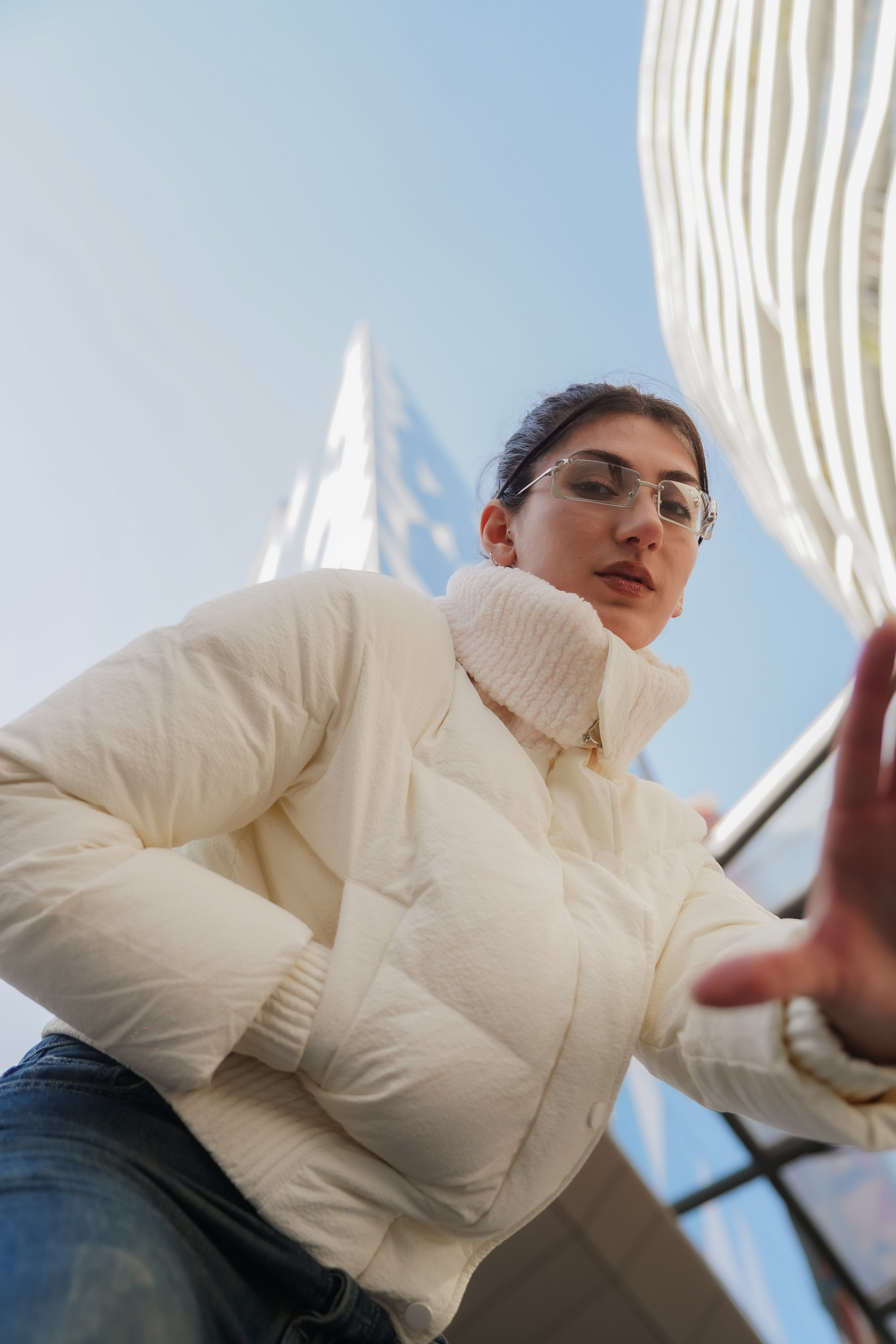 A young woman wearing glasses and a white jacket, taking a selfie outdoors with modern white buildings and a clear blue sky in the background.