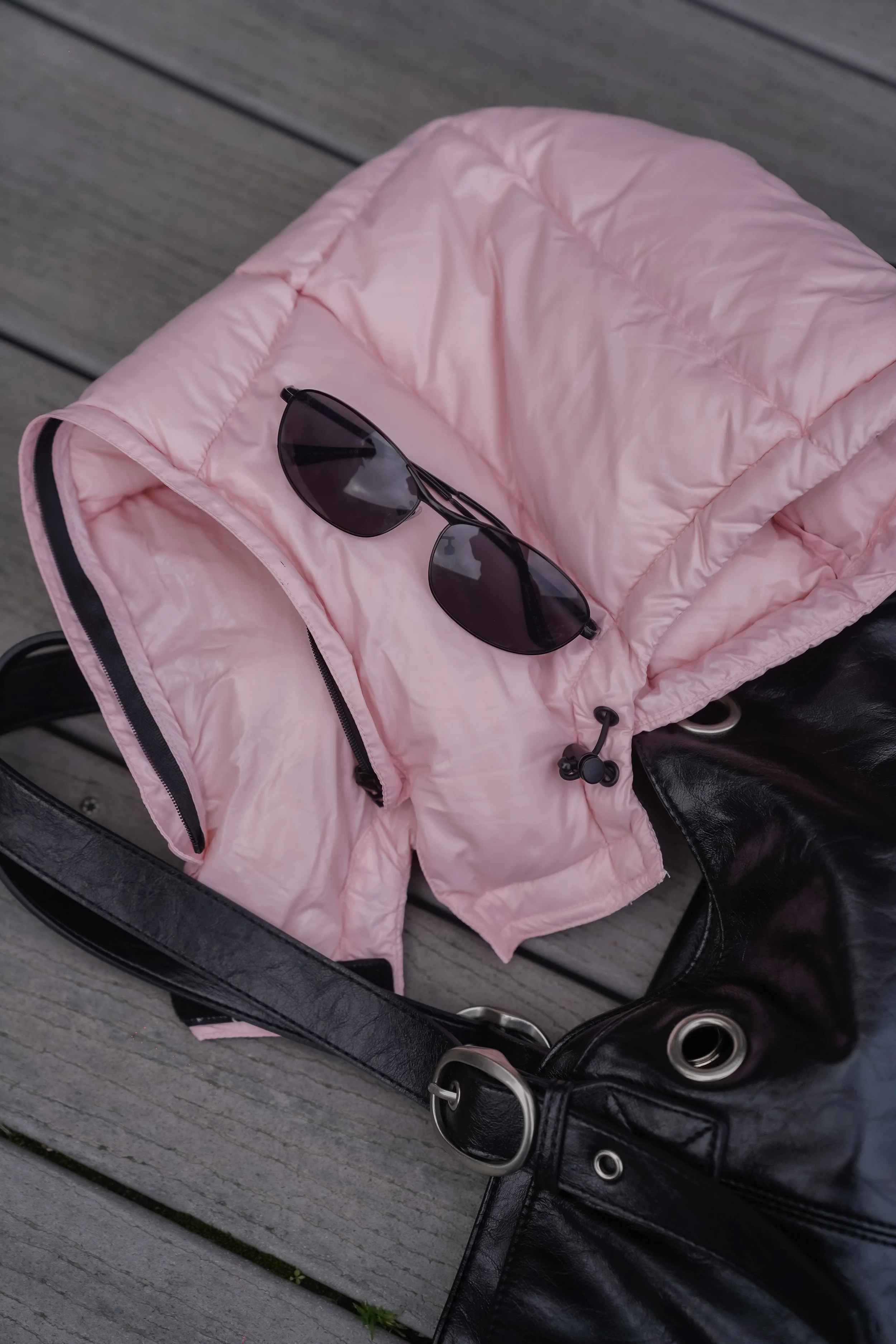 Pink puffer jacket with sunglasses resting on top, placed on a black chair on a wooden surface.
