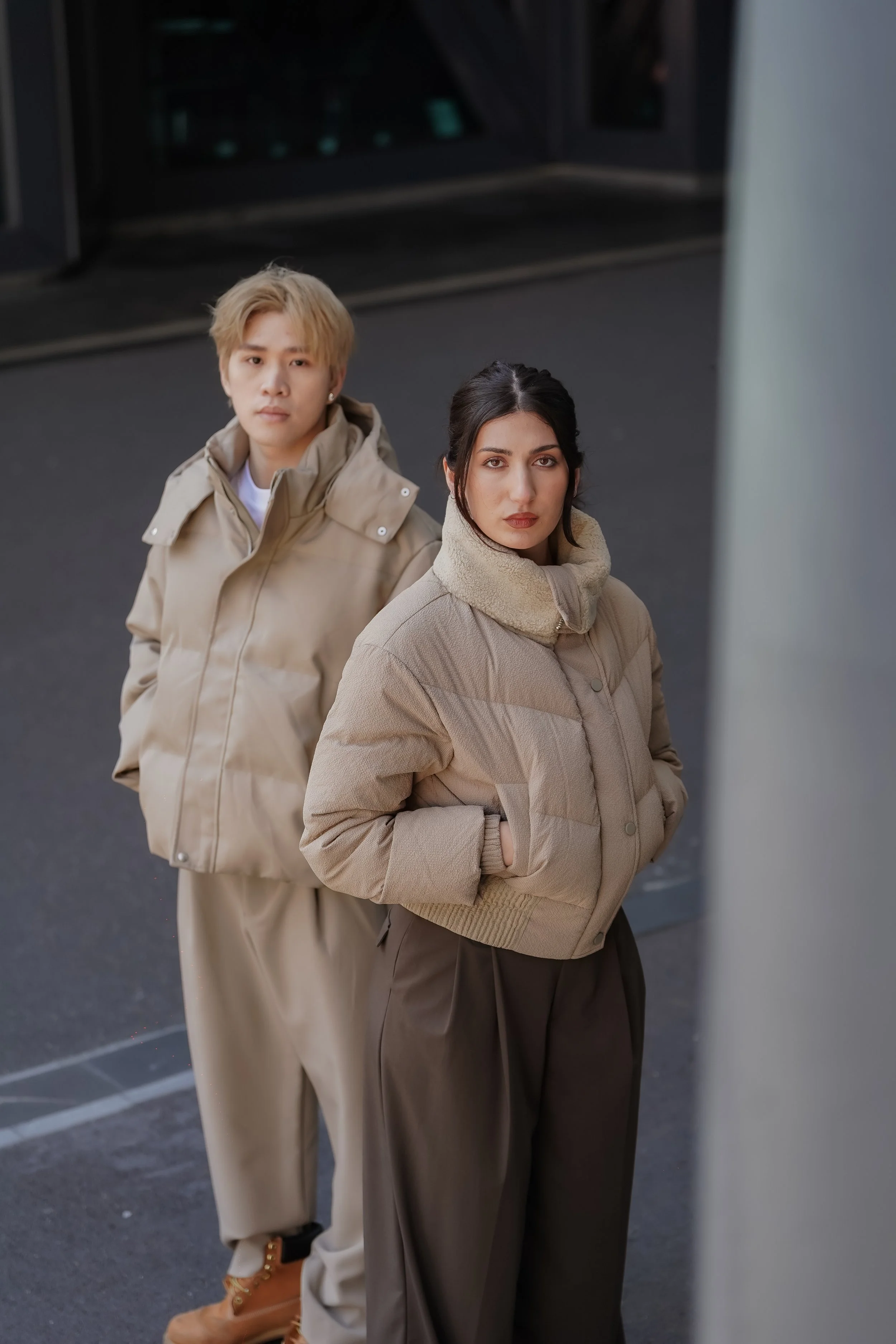Two women standing outdoors against a dark background, wearing beige and brown warm clothing, looking directly at the camera.