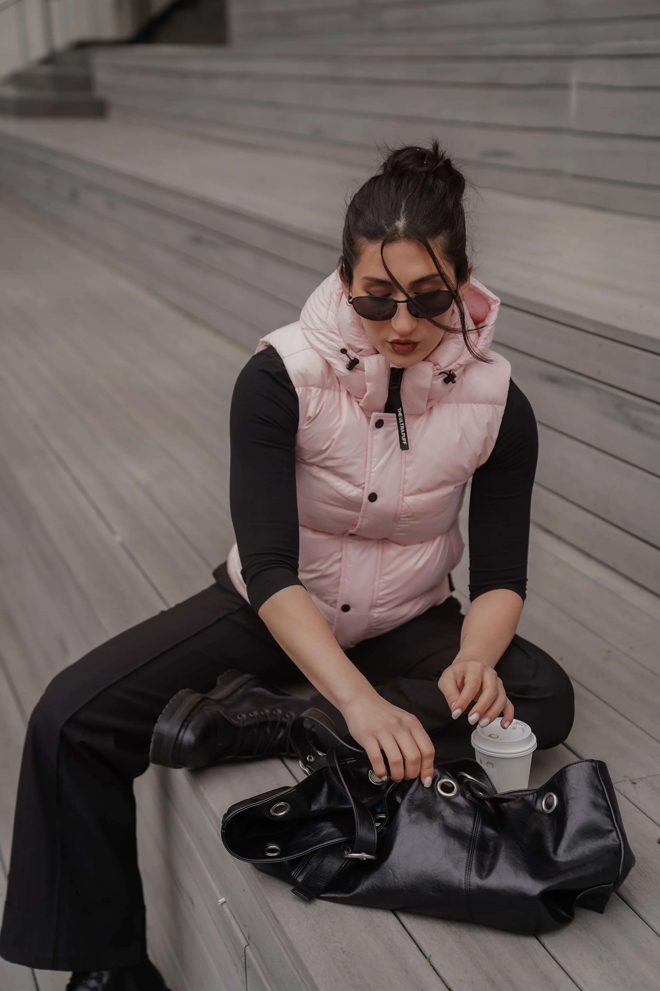 A woman sitting on a bench, wearing sunglasses, a pink padded vest, and black pants, pouring from her black leather bag and holding a coffee cup, with a large bag and backpack nearby.