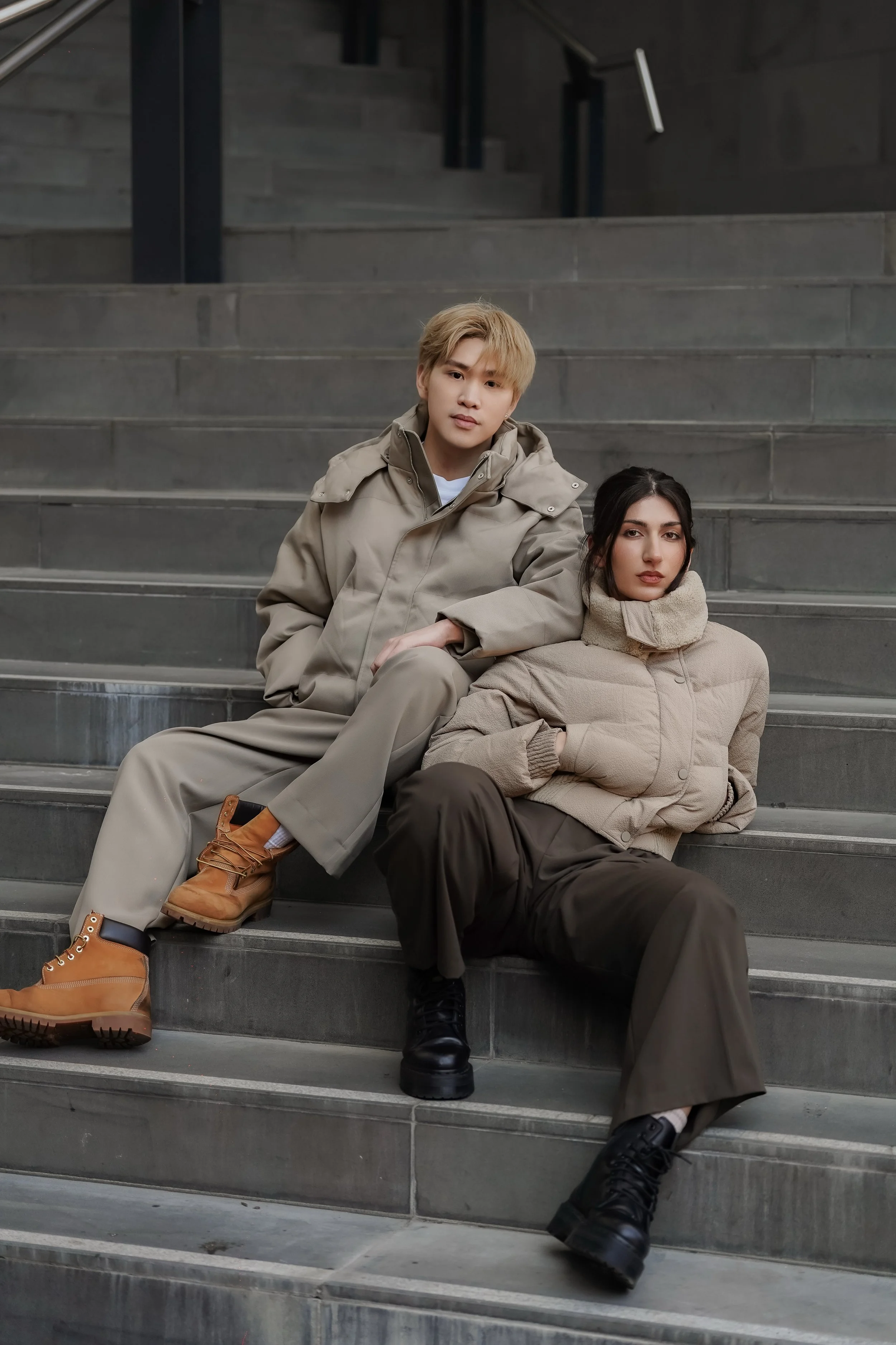 Two young adults sitting on concrete stairs outdoors. The person on the left is a blonde-haired man wearing a beige coat, beige pants, and tan boots. The person on the right is a dark-haired woman wearing a beige coat, dark pants, and black shoes.