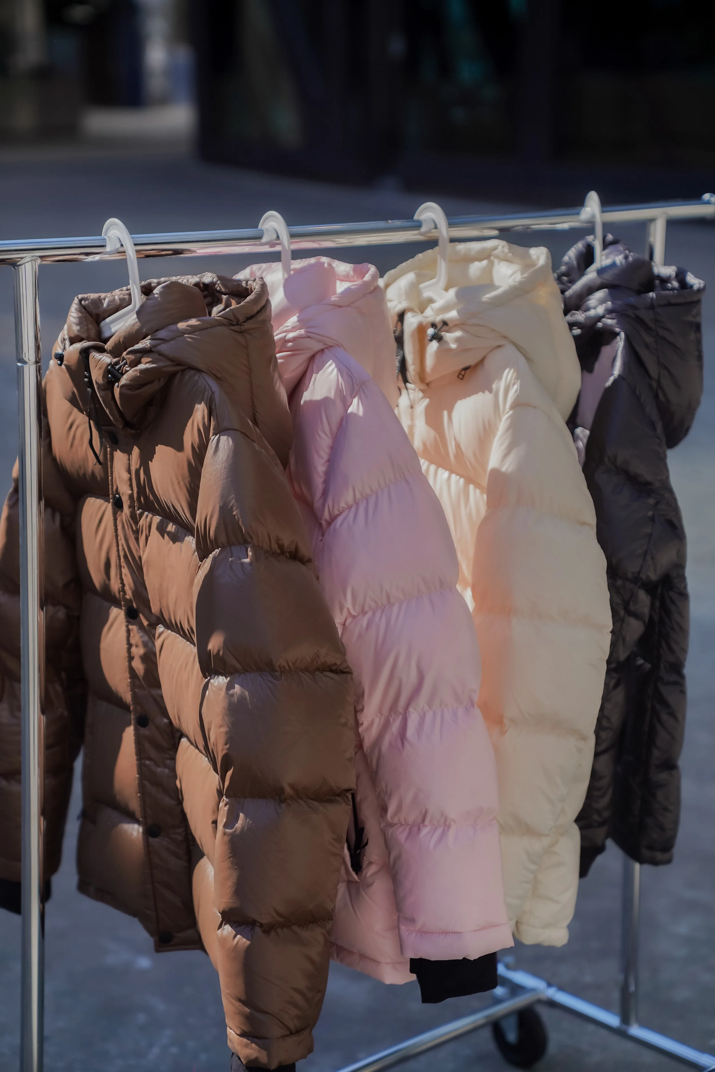 A metal clothing rack with several puffy winter jackets hanging on white plastic hangers, set outdoors in a shaded area.