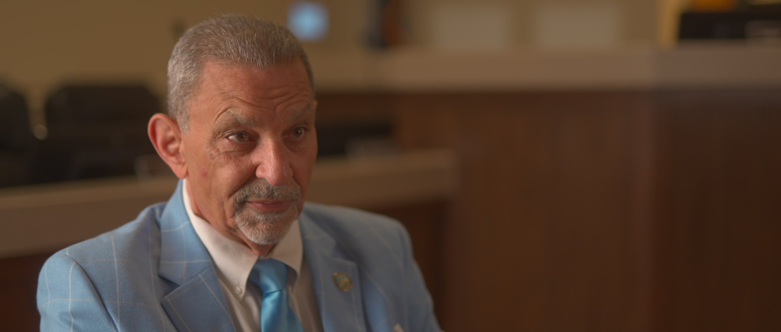 Close-up image of an older man with gray hair and beard, wearing a light blue suit and tie, sitting indoors.
