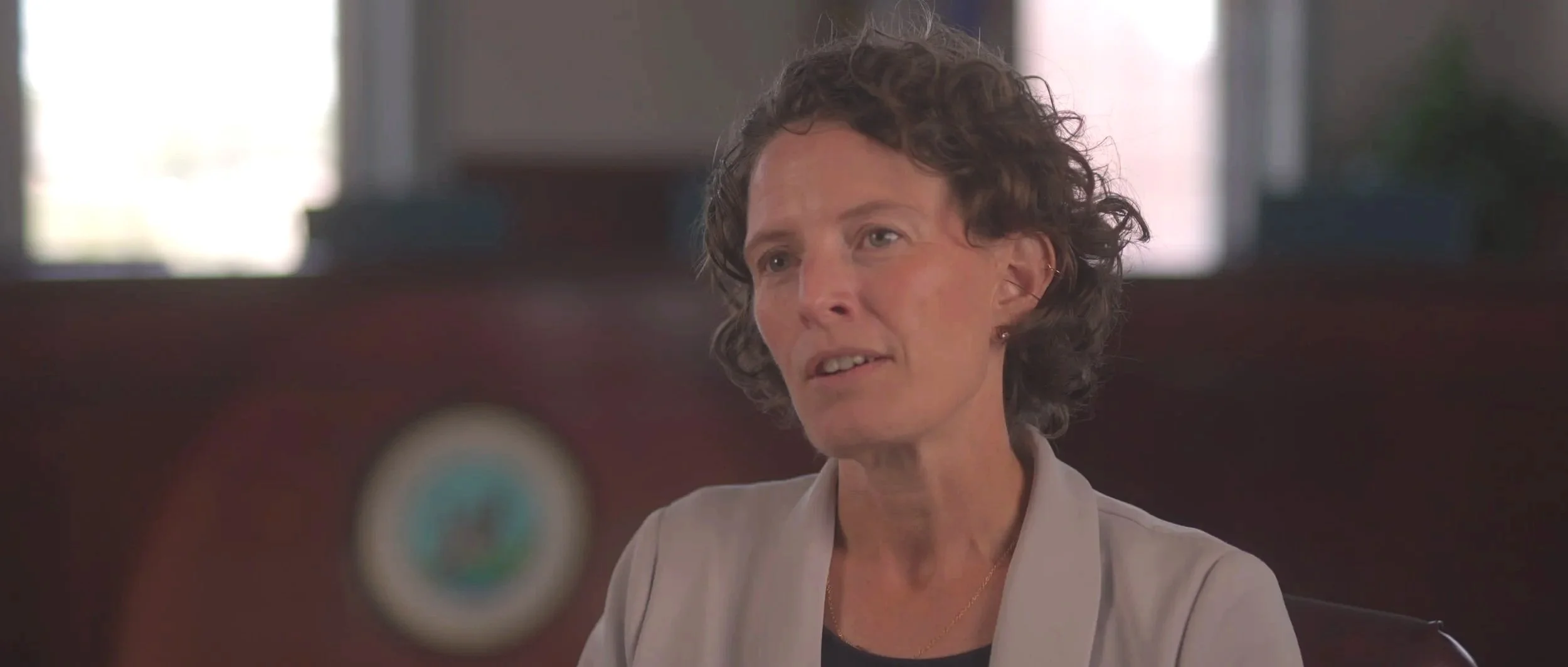 A woman with short, curly gray hair and a light-colored blazer sitting indoors, speaking or listening, with a blurred background.