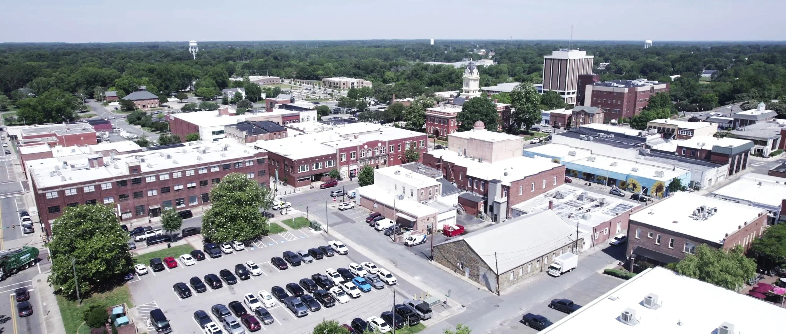 Aerial view of a small downtown area with brick buildings, parking lots, and trees, with a large forested area in the background.