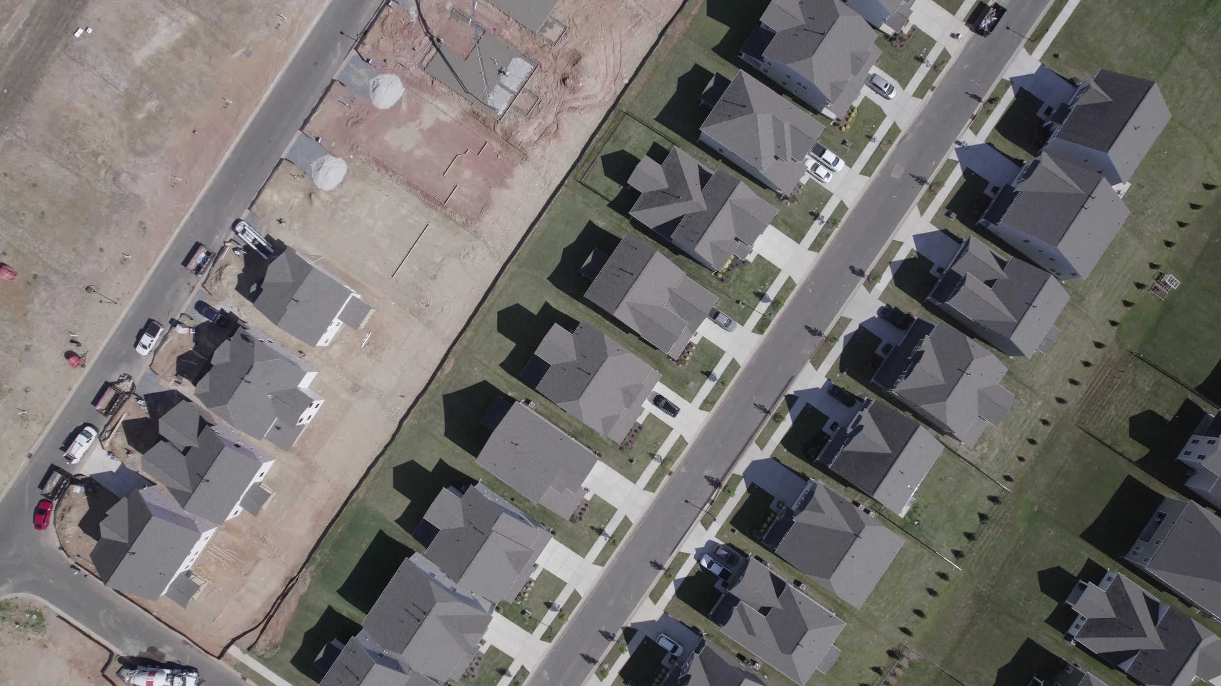 An aerial view of a suburban neighborhood showing houses, cars parked along the street, and a construction site with dug-up ground and building foundations.