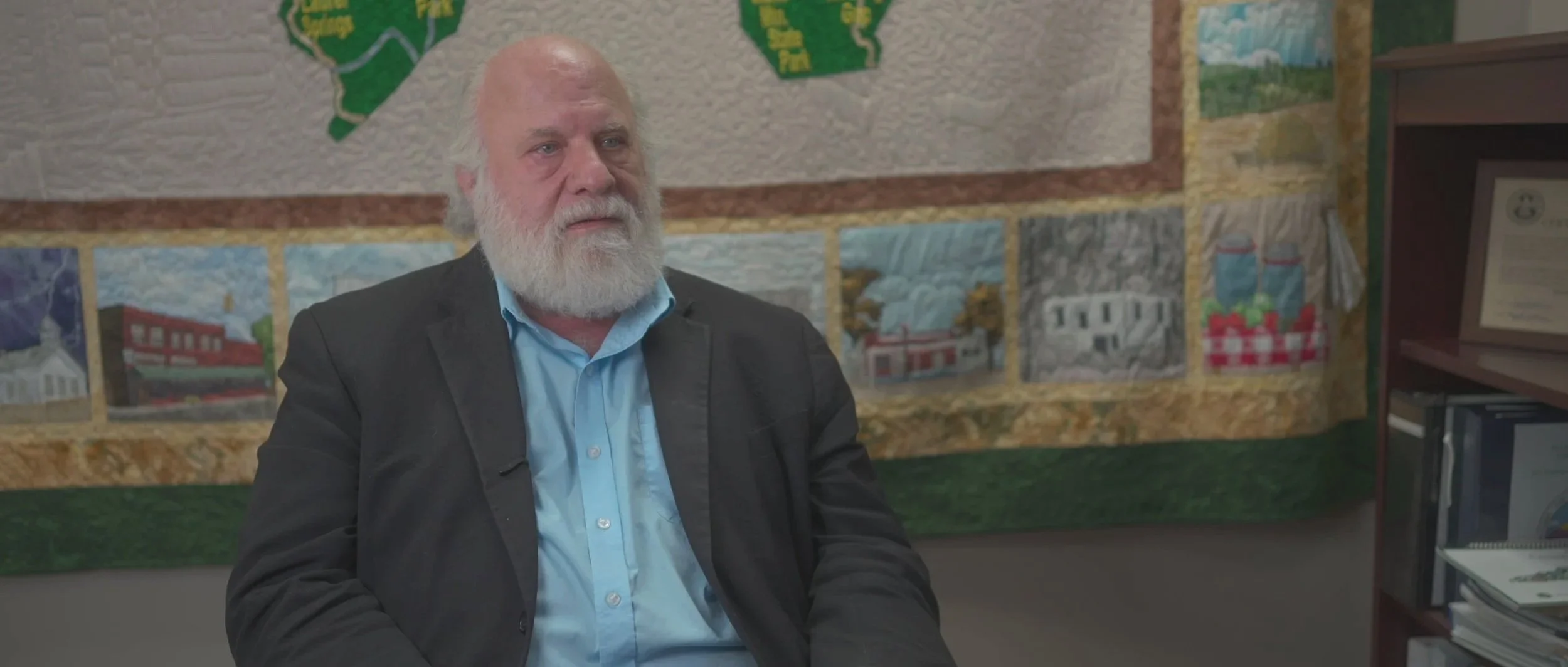 An older man with a white beard and blonde hair, wearing a black jacket and light blue shirt, sitting in front of a colorful quilted wall hanging depicting various buildings and landscapes.