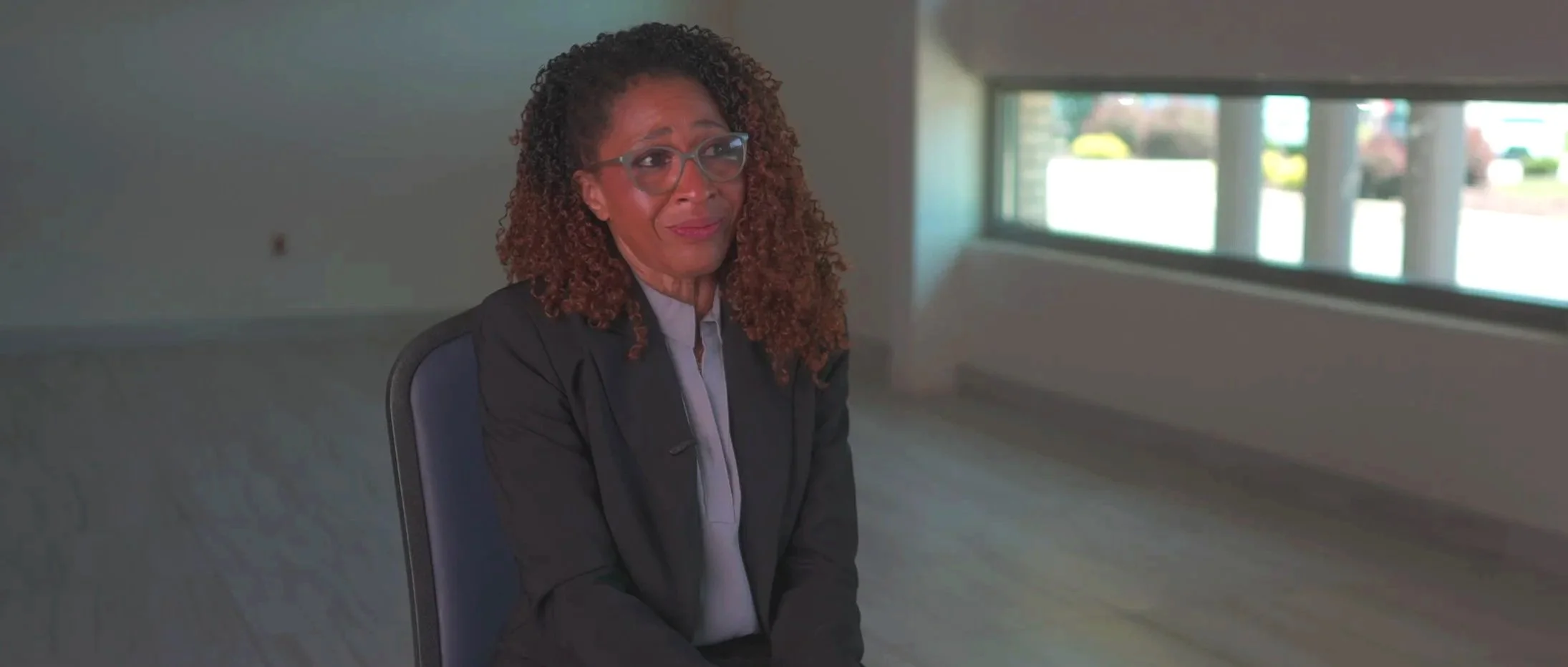 A woman with curly hair, glasses, and a black jacket sitting on a chair in an indoor setting with large windows.
