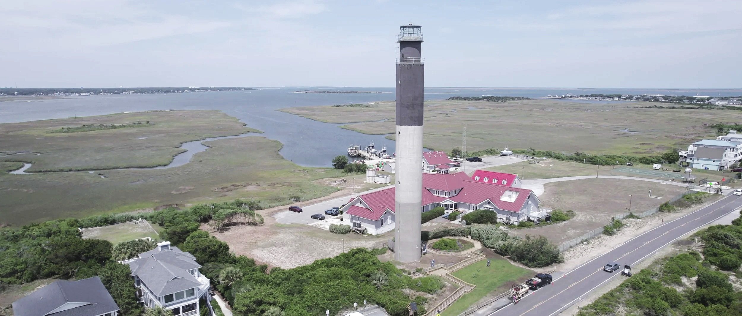 A tall lighthouse near the coast with houses and green vegetation around, overlooking marshlands and water bodies.