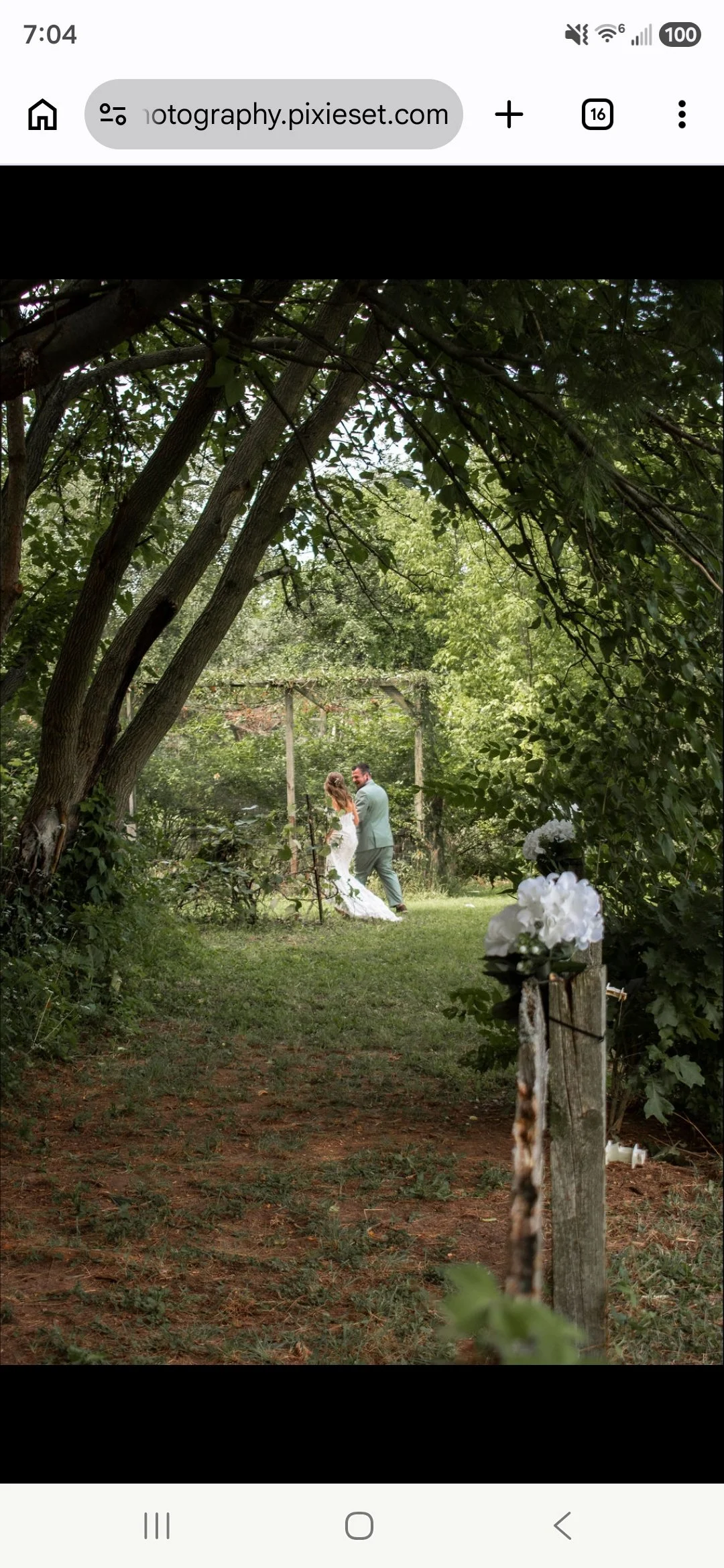 A bride and groom standing beneath trees in a garden setting, likely during a wedding ceremony, with natural greenery and a rustic wooden arch in the background.