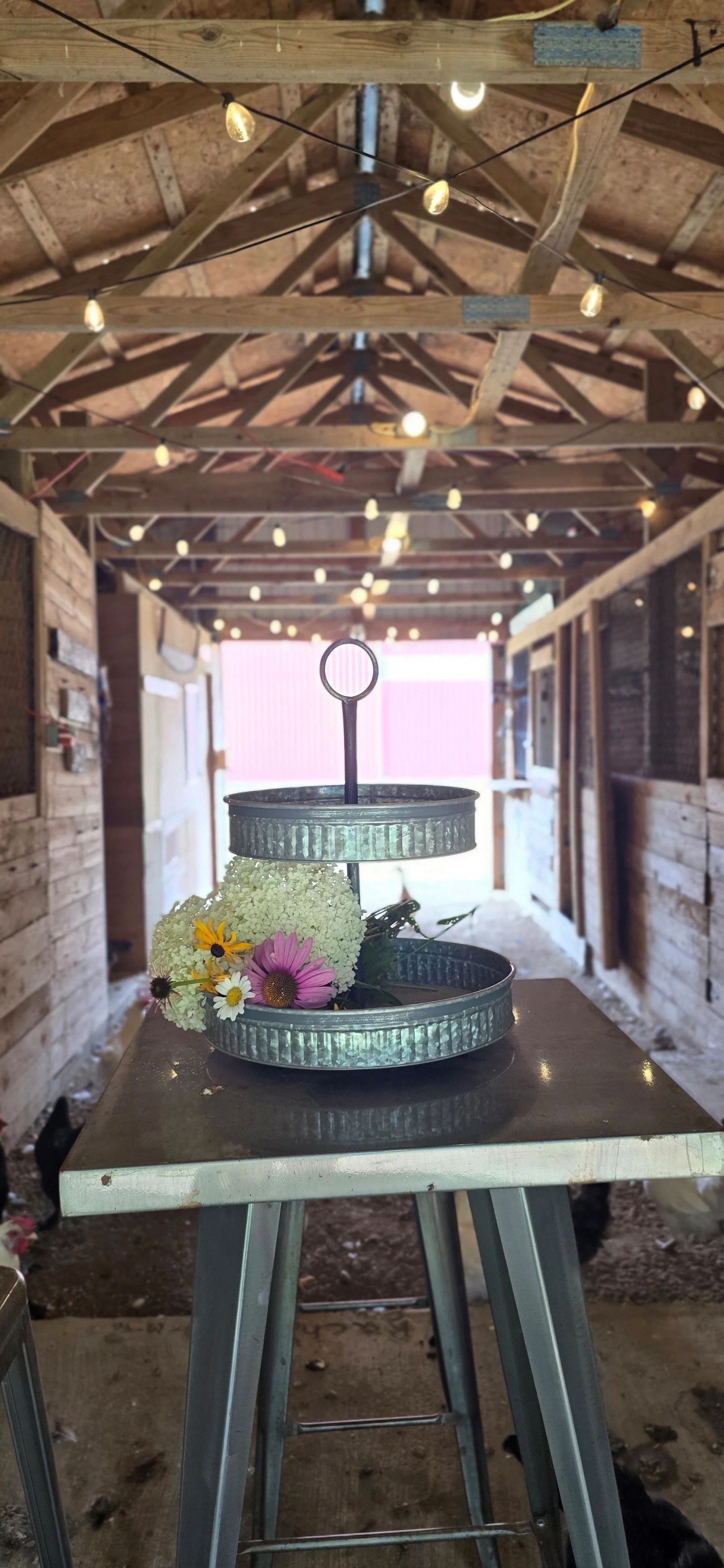 A metal two-tiered serving tray with a floral arrangement of white, yellow, and pink flowers on a dark table inside a barn with exposed wood rafters and string lights overhead.