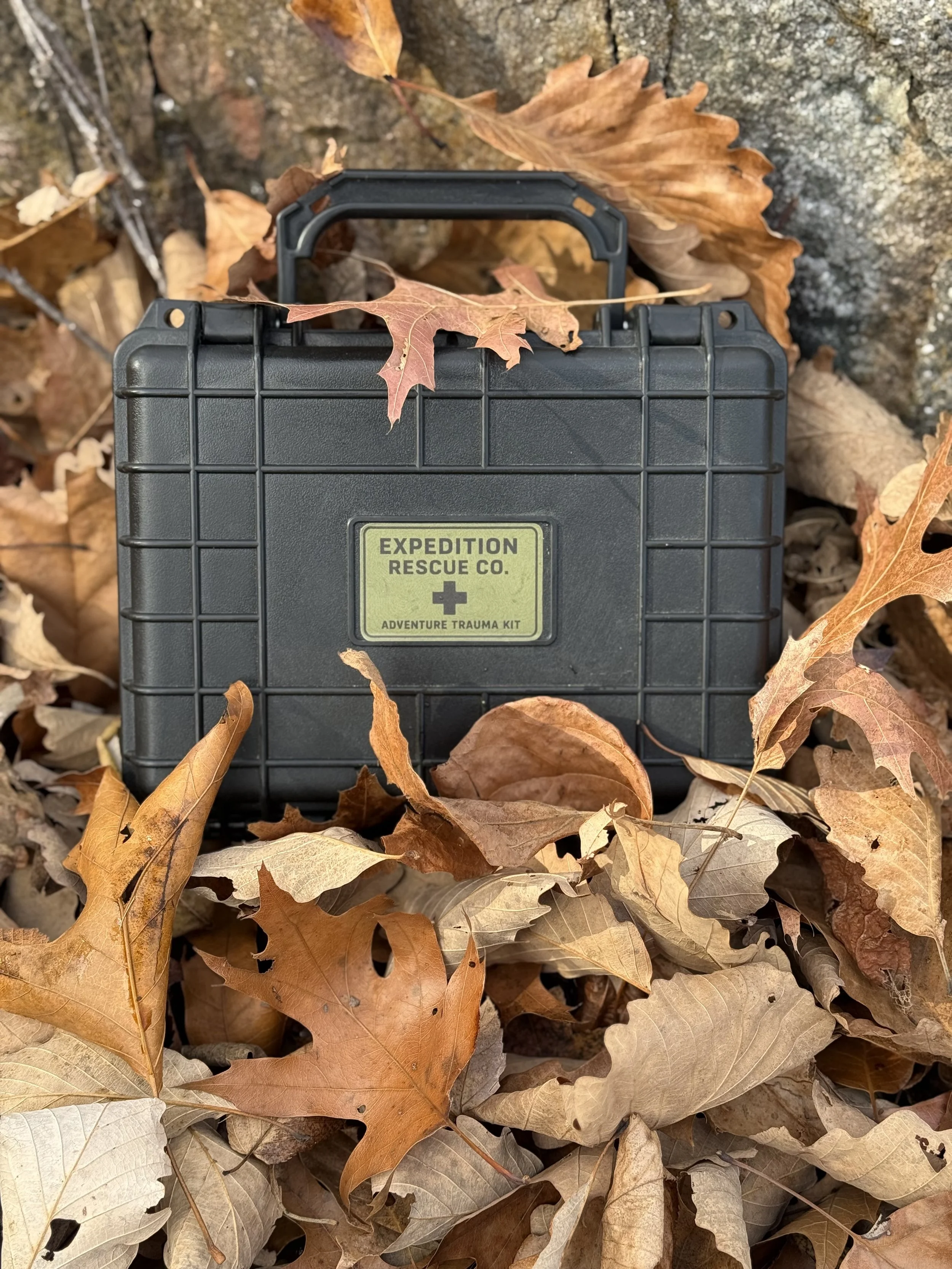 A black expedition rescue kit box on the ground surrounded by fallen autumn leaves in shades of brown, tan, and orange.
