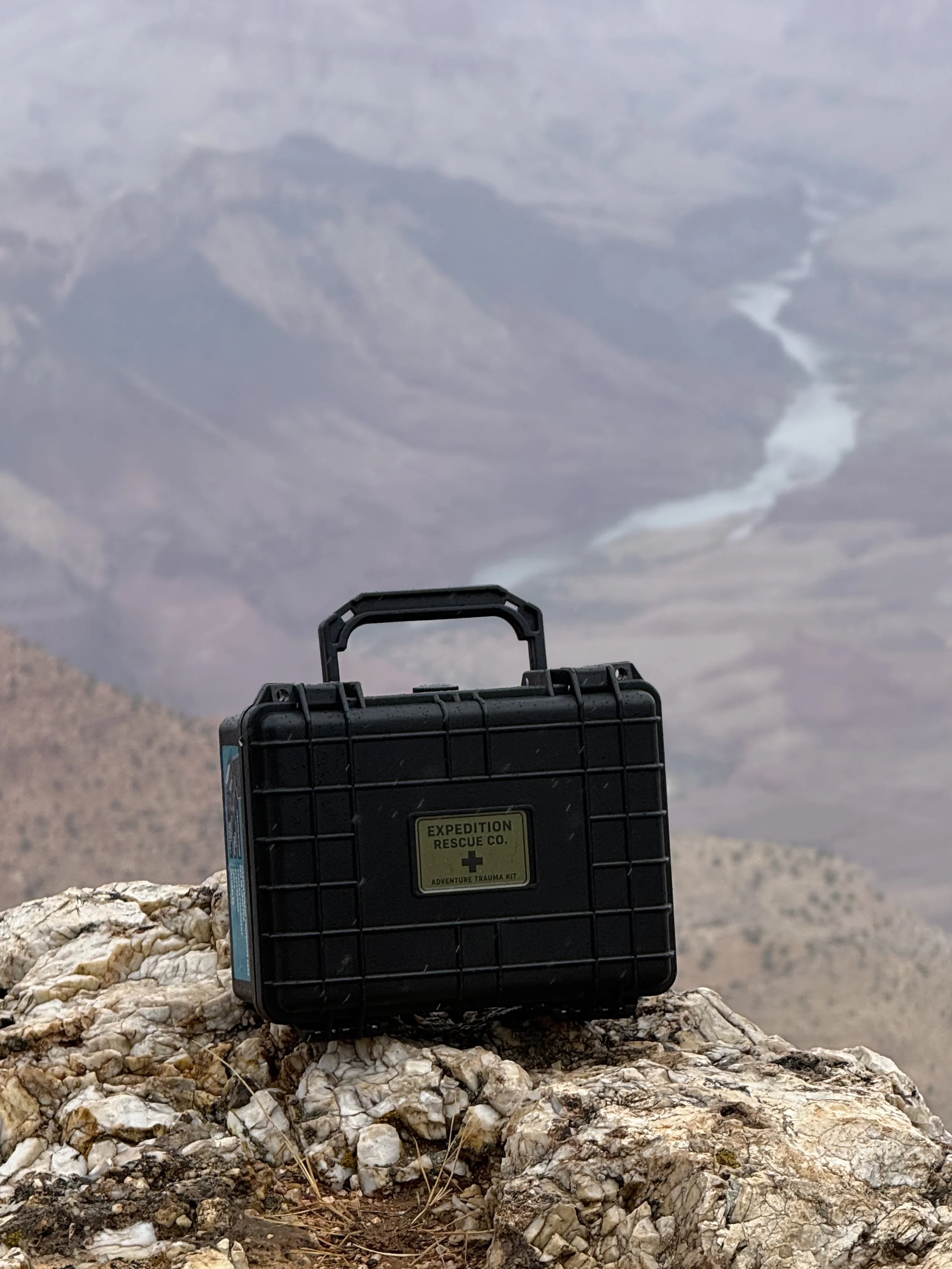 A black adventure trauma kit box from Expedition Rescue Co. sits on a rocky surface in a desert landscape with mountains in the background and a cloudy sky overhead.
