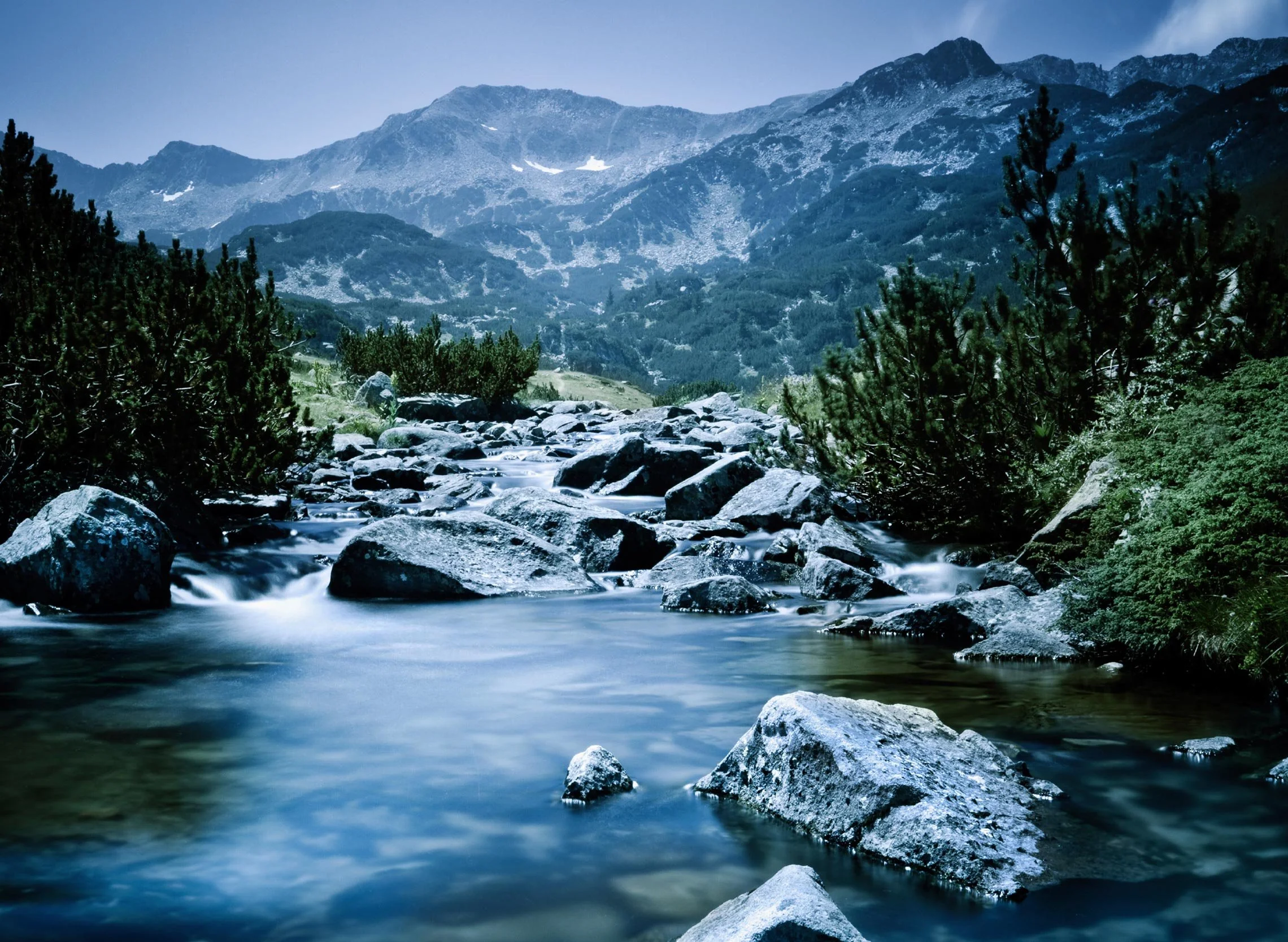 A mountain landscape with tall rocky peaks and patches of snow, dense green trees in the foreground, and a flowing river at the base.