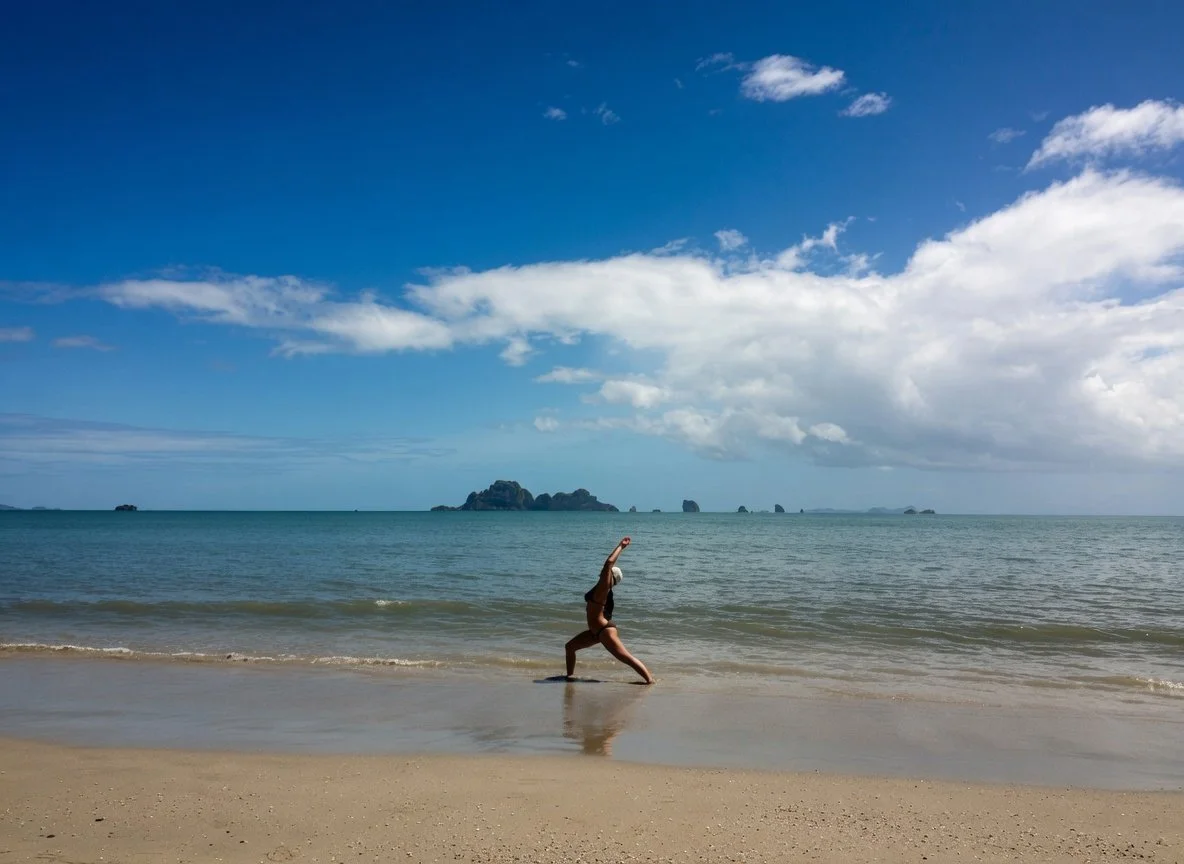 A woman performing a handstand on the beach close to the shoreline with rocks and islands visible in the background under a partly cloudy sky.