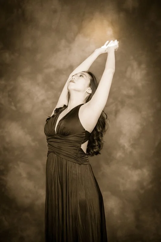 A woman in a long, dark dress with her arms raised above her head, looking upward with a serene expression.