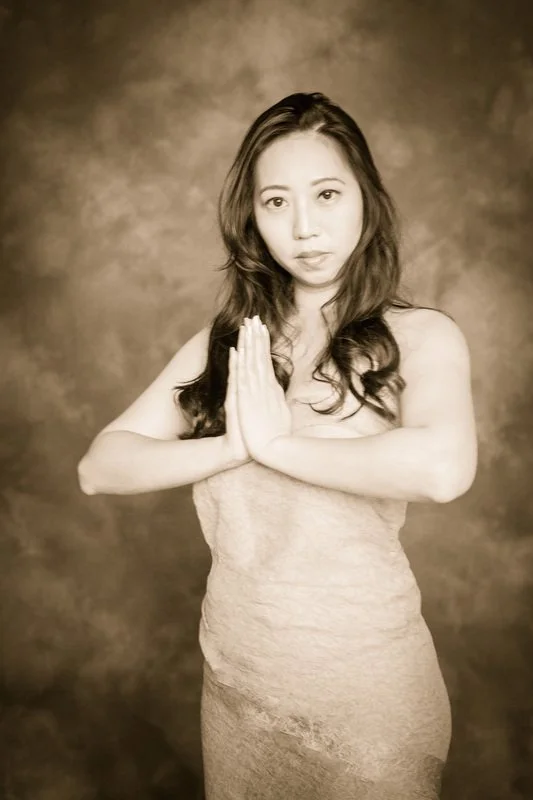 A woman with long, wavy hair, dressed in a strapless dress, standing with palms pressed together in front of her chest, looking at the camera.