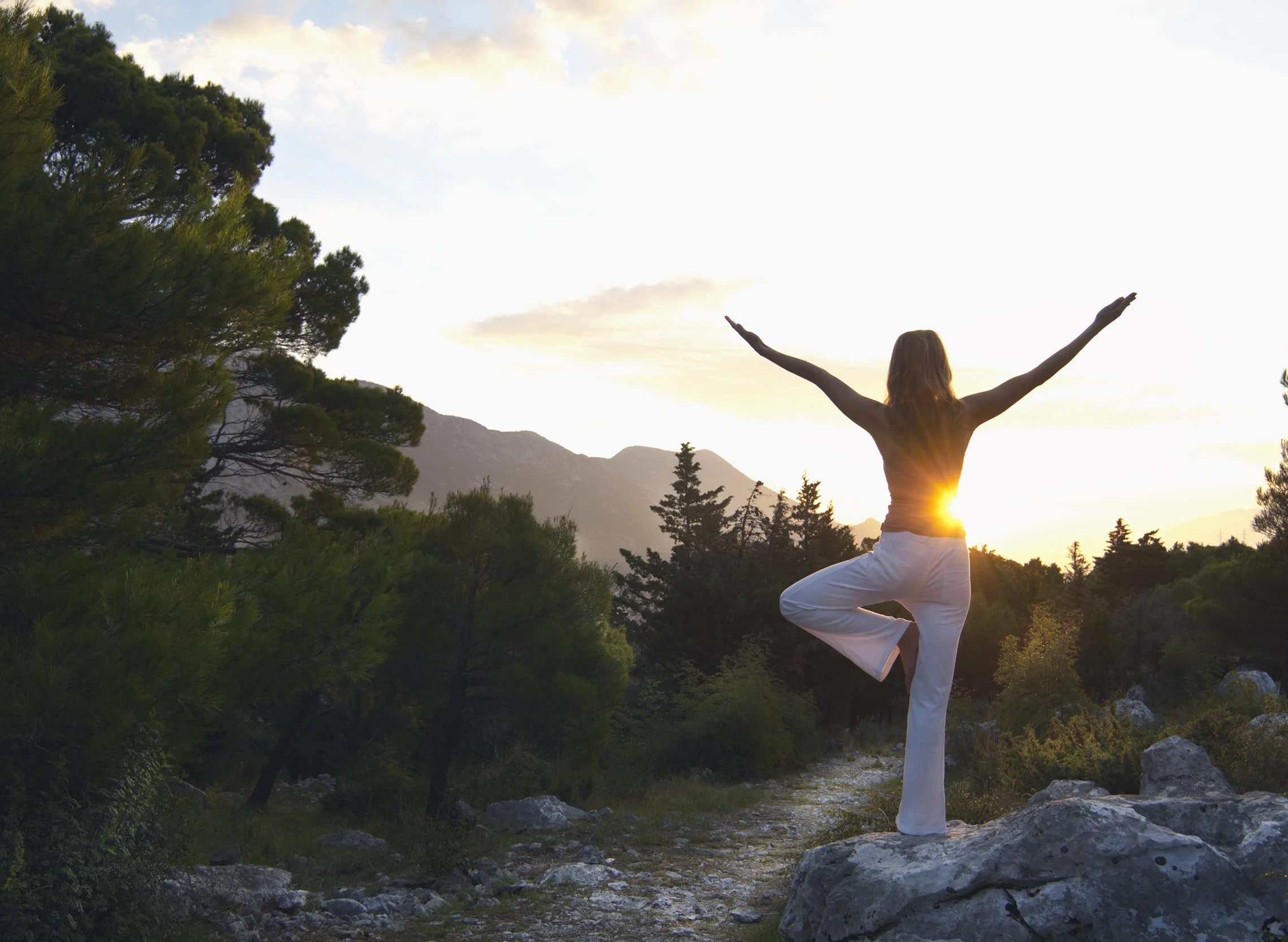 A woman practicing yoga outdoors in a mountain landscape during sunset, standing on one leg with arms stretched out wide, surrounded by trees and rocky terrain.