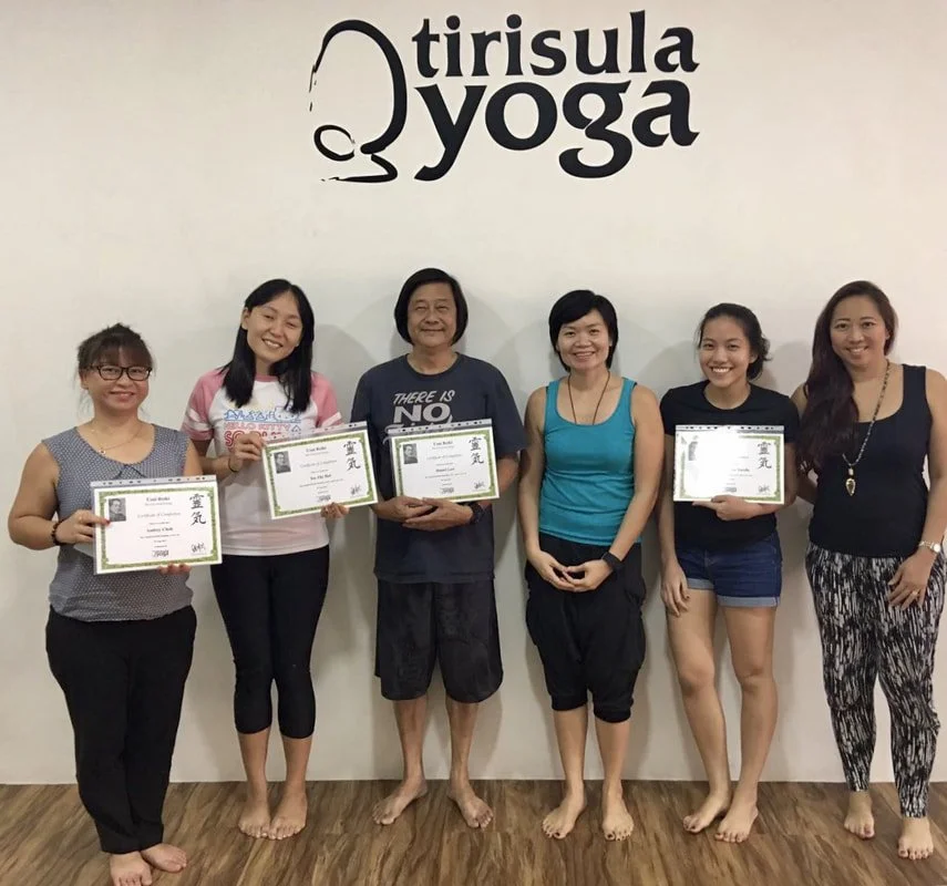 Six women standing in front of a wall with the Tririsula Yoga logo, holding certificates and smiling. They are in a yoga studio with wooden flooring.