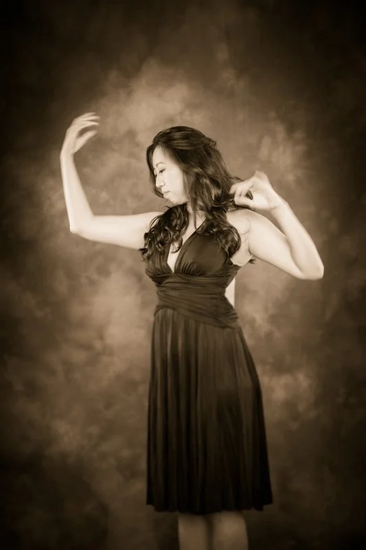 A woman in a dark sleeveless dress with long flowing hair poses with her eyes closed against a textured brown backdrop.