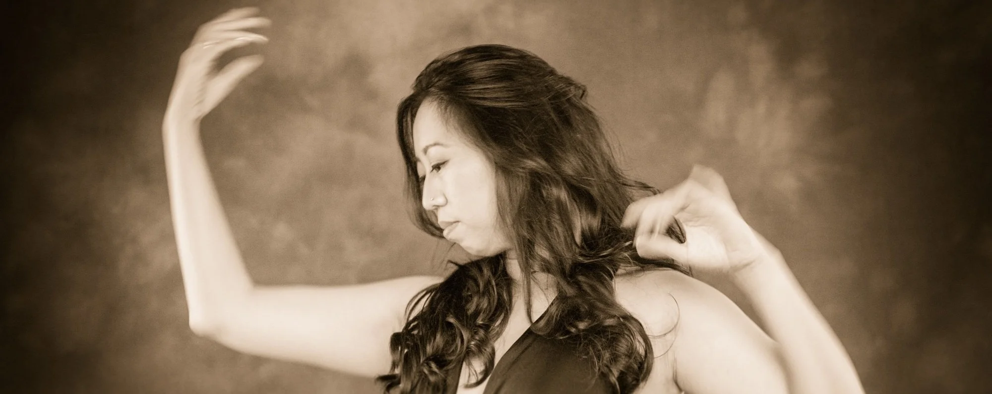 Sepia-toned photo of a woman with long wavy hair, her eyes closed, raising her arms with her hands near her head, wearing a sleeveless top.
