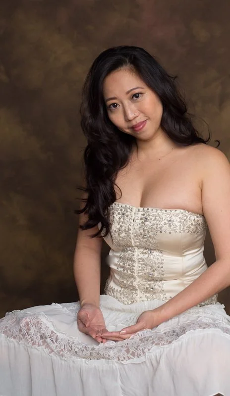 A young woman with long dark hair and a light complexion, wearing a strapless cream-colored dress with intricate embroidery, sitting with her hands folded on her lap, against a brown mottled background.
