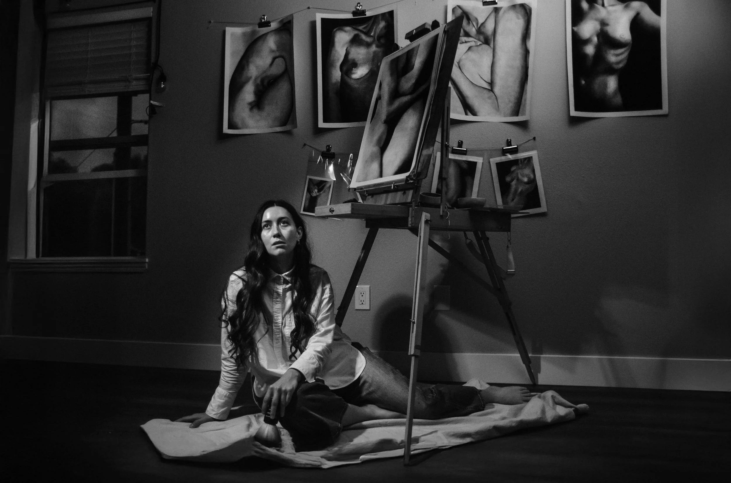 Black and white photo of a woman with long hair sitting on the floor in front of a wall decorated with drawings of human torsos. She is wearing a light-colored shirt and dark pants.