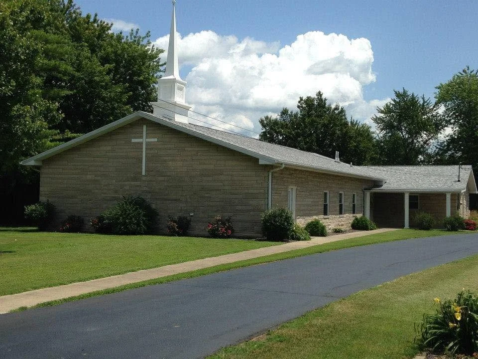 A brick church with a white steeple, green lawn, and landscaped bushes, under a partly cloudy sky.