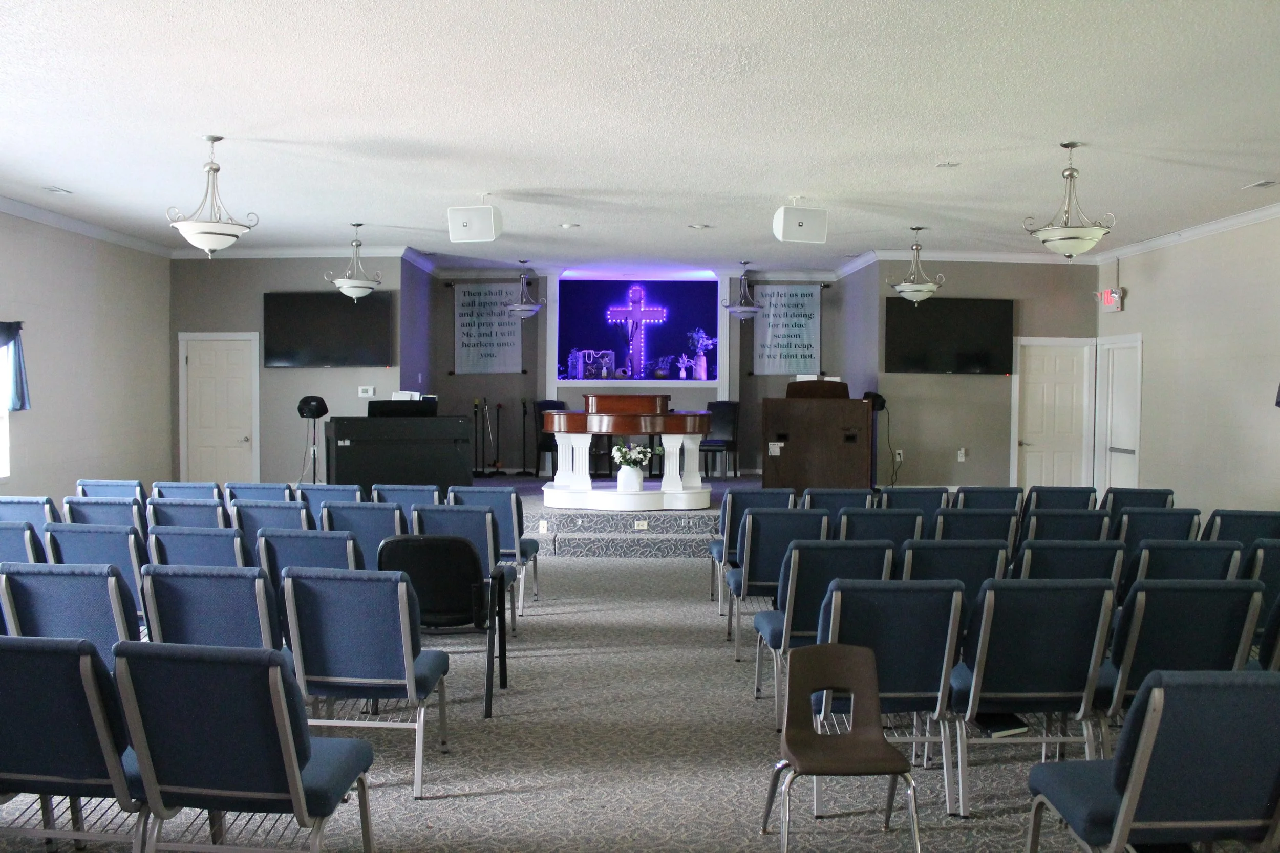 Interior of a church auditorium with blue rows of chairs, a stage with a white pulpit, a cross lit with purple neon lights, and two banners with scripture verses on either side of the cross.
