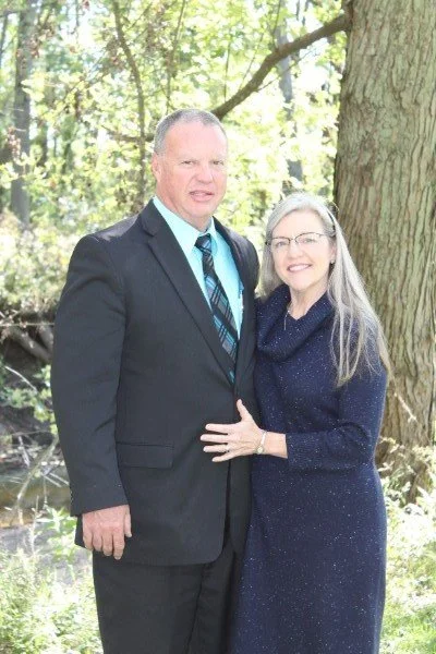 A man in a suit and a woman in a dark dress standing outdoors near a large tree, smiling at the camera.