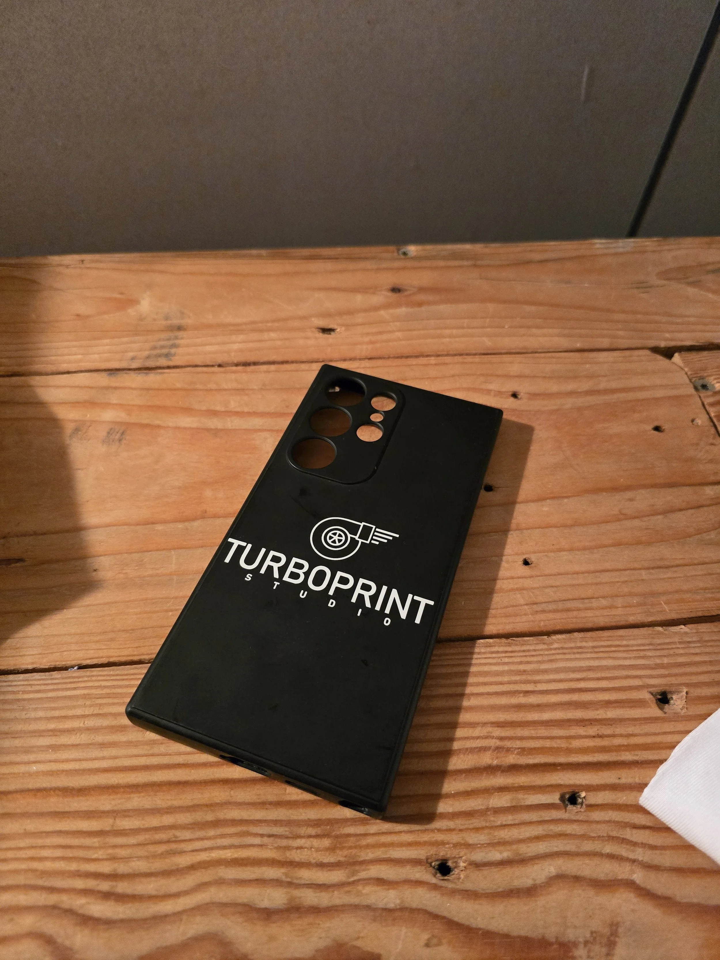 Close-up of a black TurboPrint Studio sticker on a wooden surface, with a gray wall in the background.